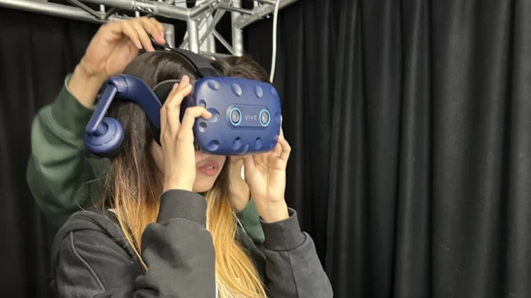 Postgraduate student putting on VR headset with help of another in a VR booth